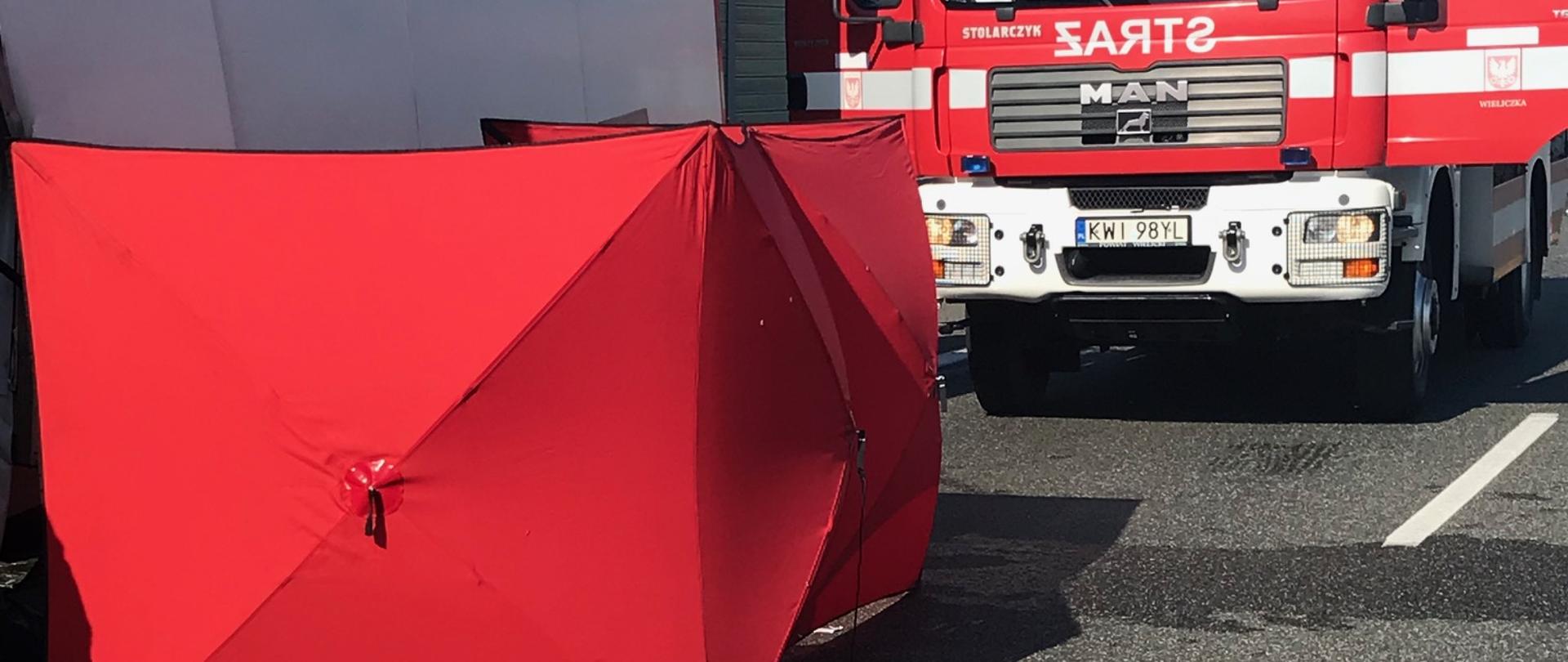 Na zdjęciu widoczny parawan osłaniający miejsce prowadzonych działań. Z za parawanu wyłania się samochód dostawczy. Na drugim planie znajduje się samochód pożarniczy.