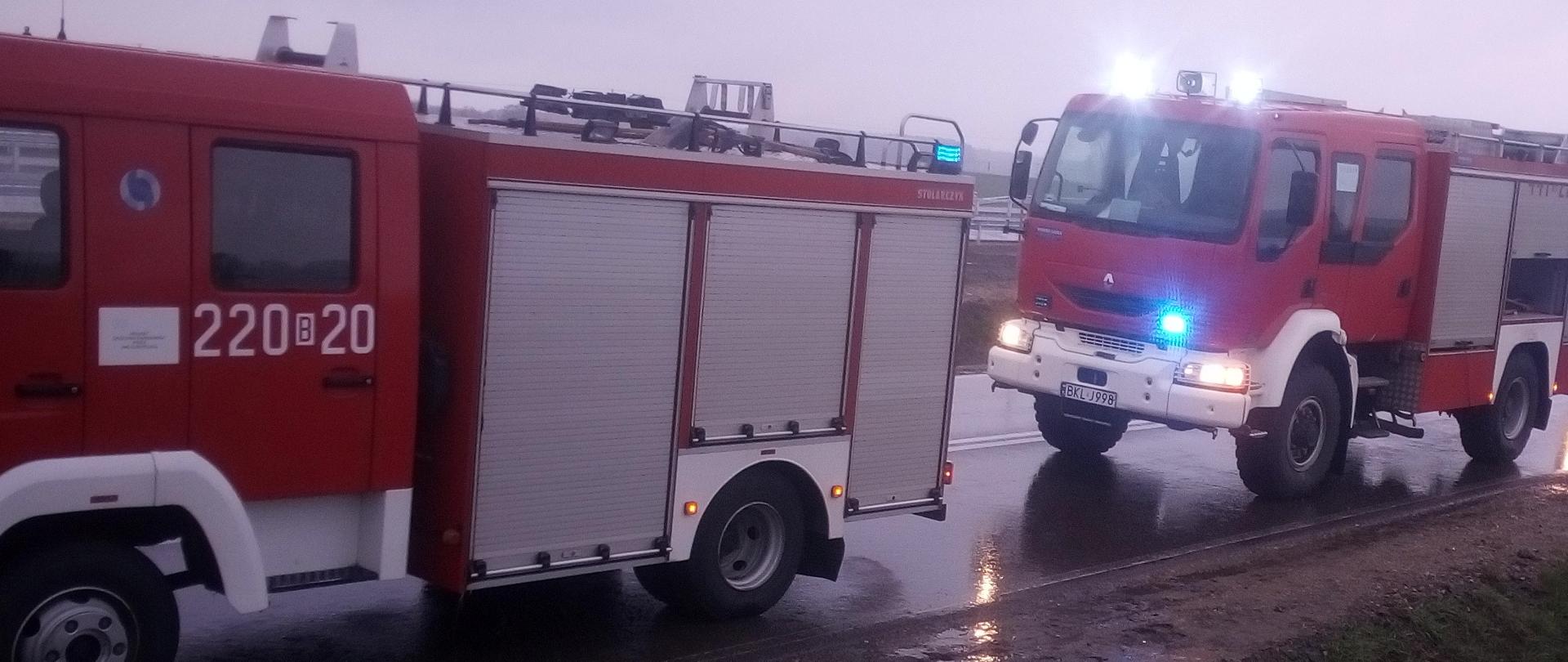Zdjęcie przedstawia dwa samochody pożarnicze stojące na poboczu drogi z włączonymi światłami błyskowymi.