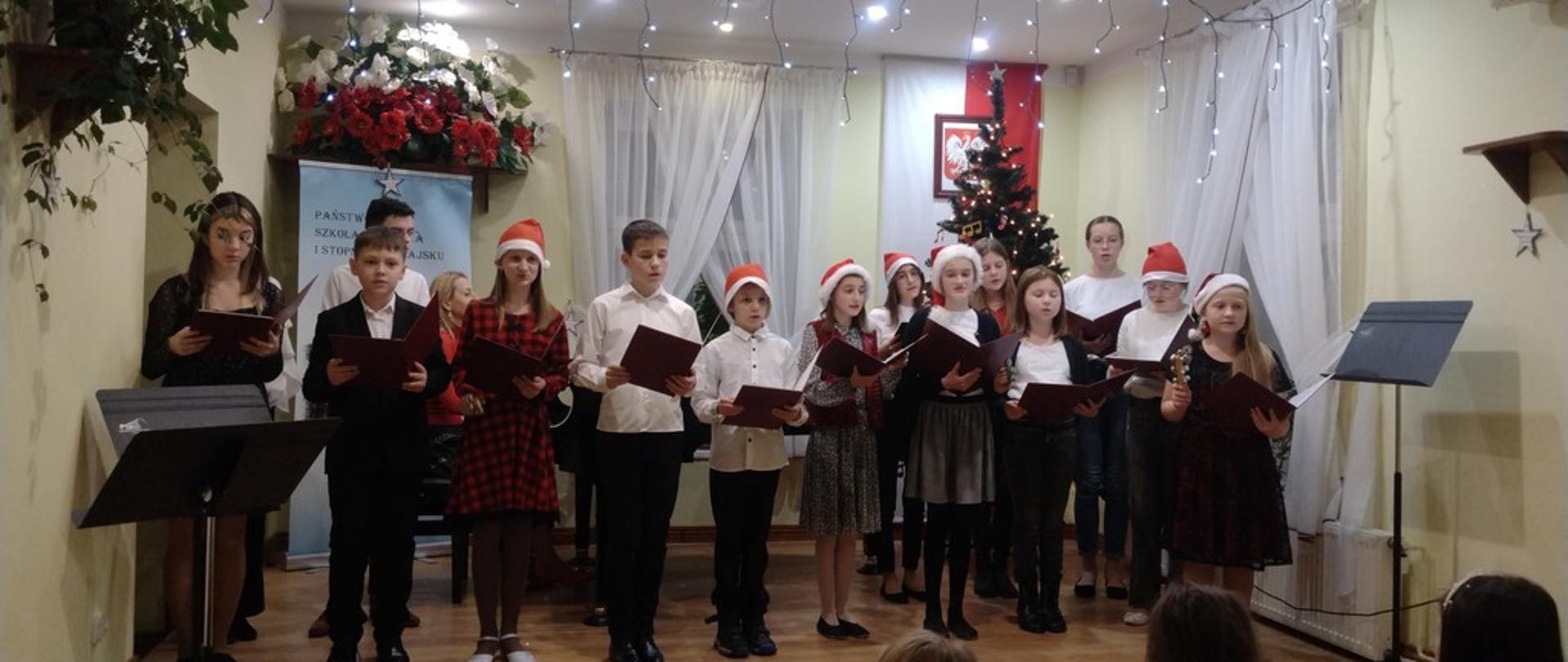 Chór szkolny na scenie w Auli PSM I st. w Leżajsku. Chór w strojach swiątecznych, część uczniów w czapkach świętego Mikołaja. Z tyłu za chórem czarny fortepian z lewej strony i przystrojona choinka z prawej strony zdjęcia.