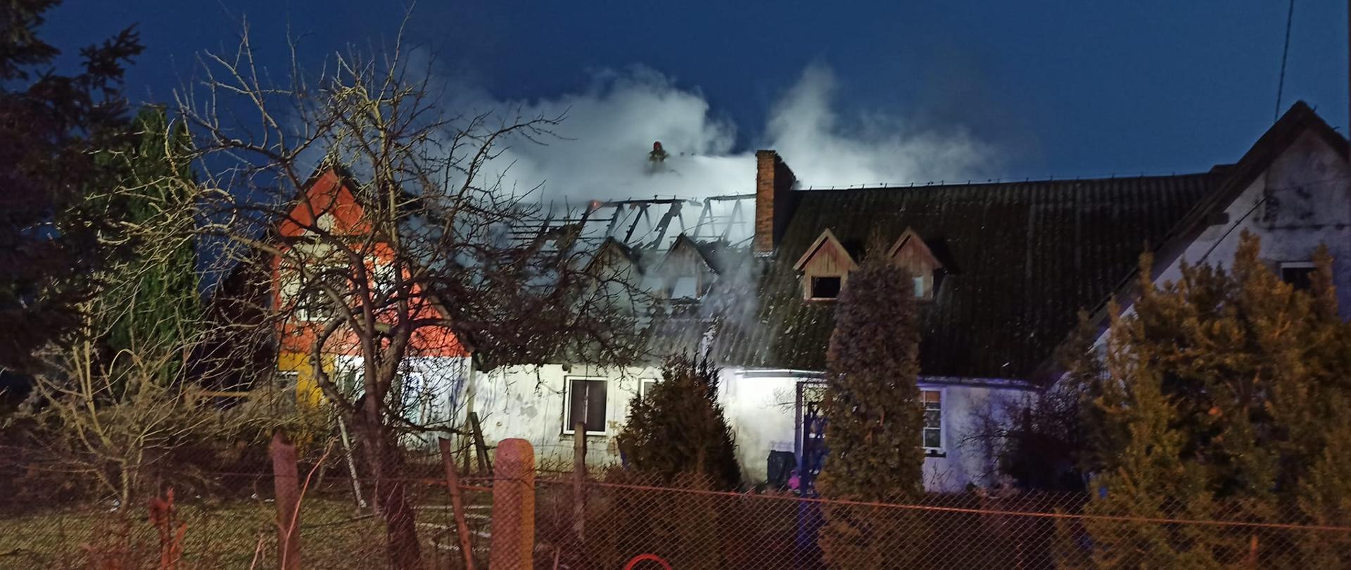 Strażacy gaszą budynek wielorodzinny widać pojazdy ratownicze, dym, mury budynku i spalona konstrukcję dachu