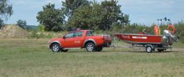 Kręci mnie bezpieczeństwo ... nad wodą. Plaża na ternie byłego poligonu wojskowego w Sierakowie. Samochód rozpoznawczo-ratowniczy ciągnie przyczepkę, na której znajduje się łódź ratownicza koloru czerwonego. W tle zarośla i drzewa. 