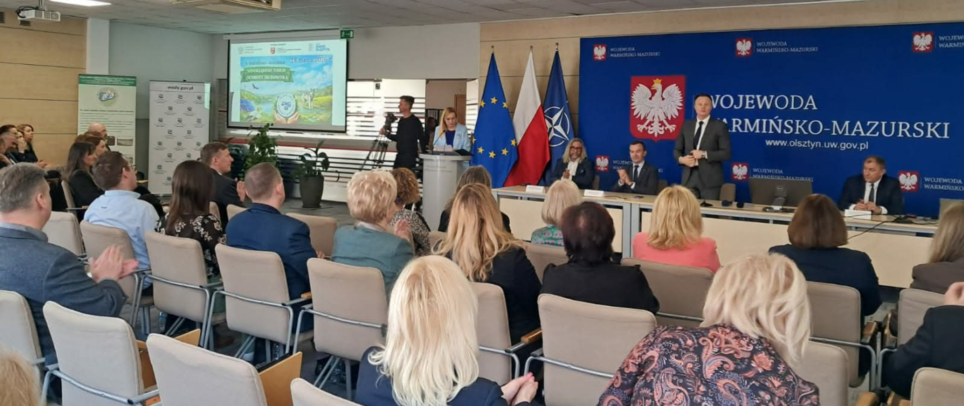 Duży stół, na którym stoją mikrofony oraz leżą dokumenty, za nim siedzą ludzie. Po lewej stronie stołu za mównicą stoi kobieta i referuje wyświetlaną prezentacje. Na przeciwko stołu siedzą ludzie i patrzą na wyświetloną prezentację. 