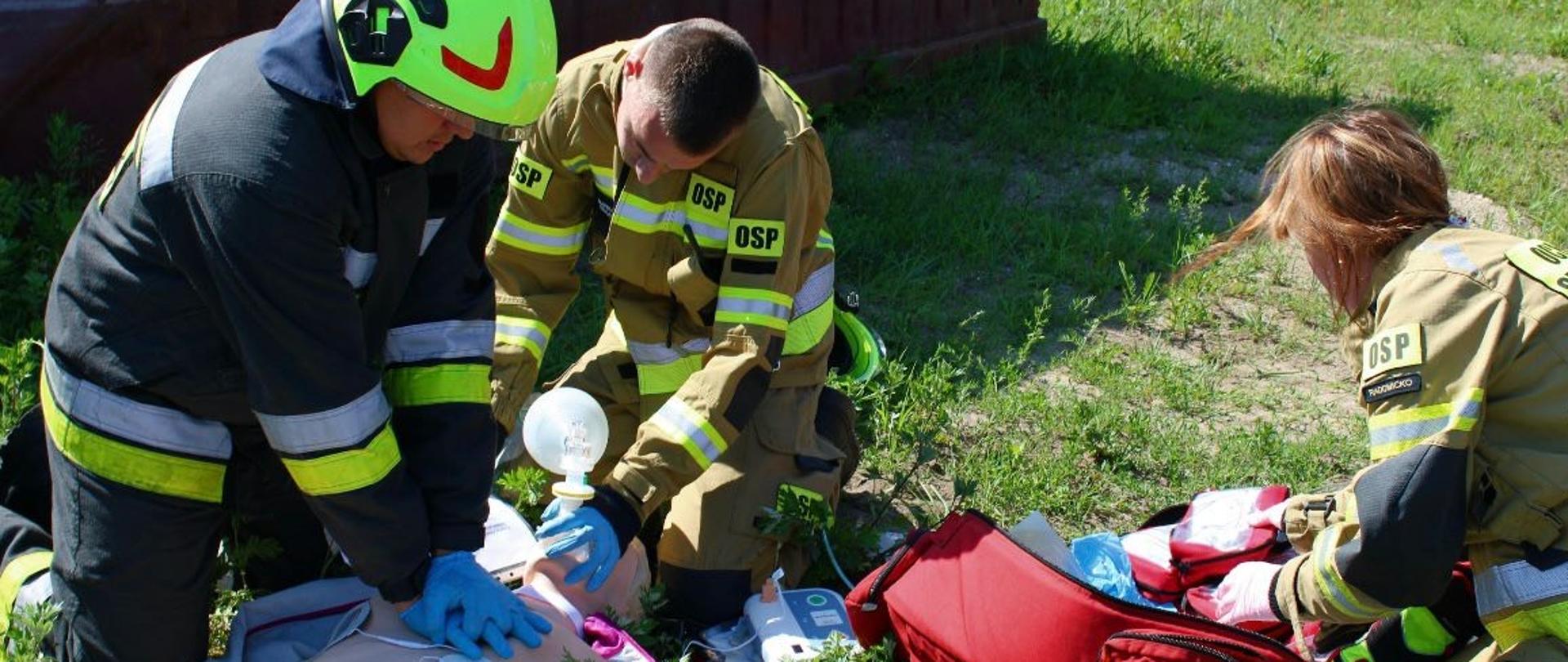 Na zdjęciu widoczni są strażacy prowadzący resuscytacje krążeniowo oddechową u poszkodowanego (fantoma) leżącego na trawie.