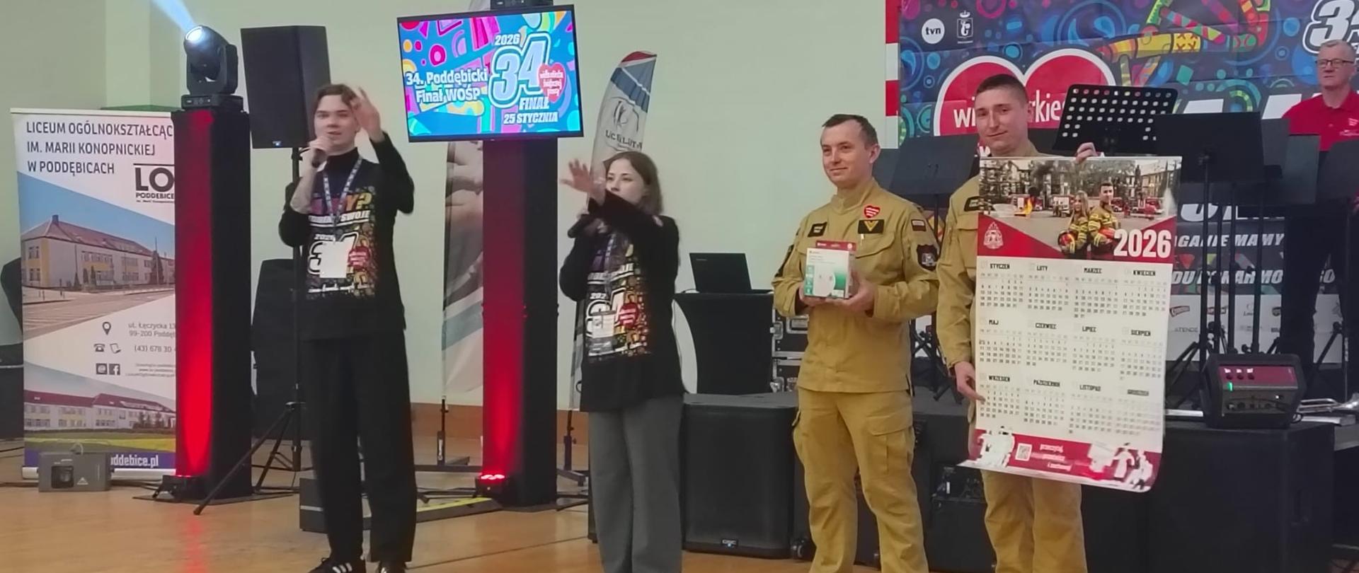 Dwóch strażaków stoi przed sceną jeden z nich trzyma kalendarz drugi czujkę tlenku węgla, obok stoją wolontariusze chłopak i dziewczyna z mikrofonami mają uniesioną rękę w górze widać że trwa licytacja
