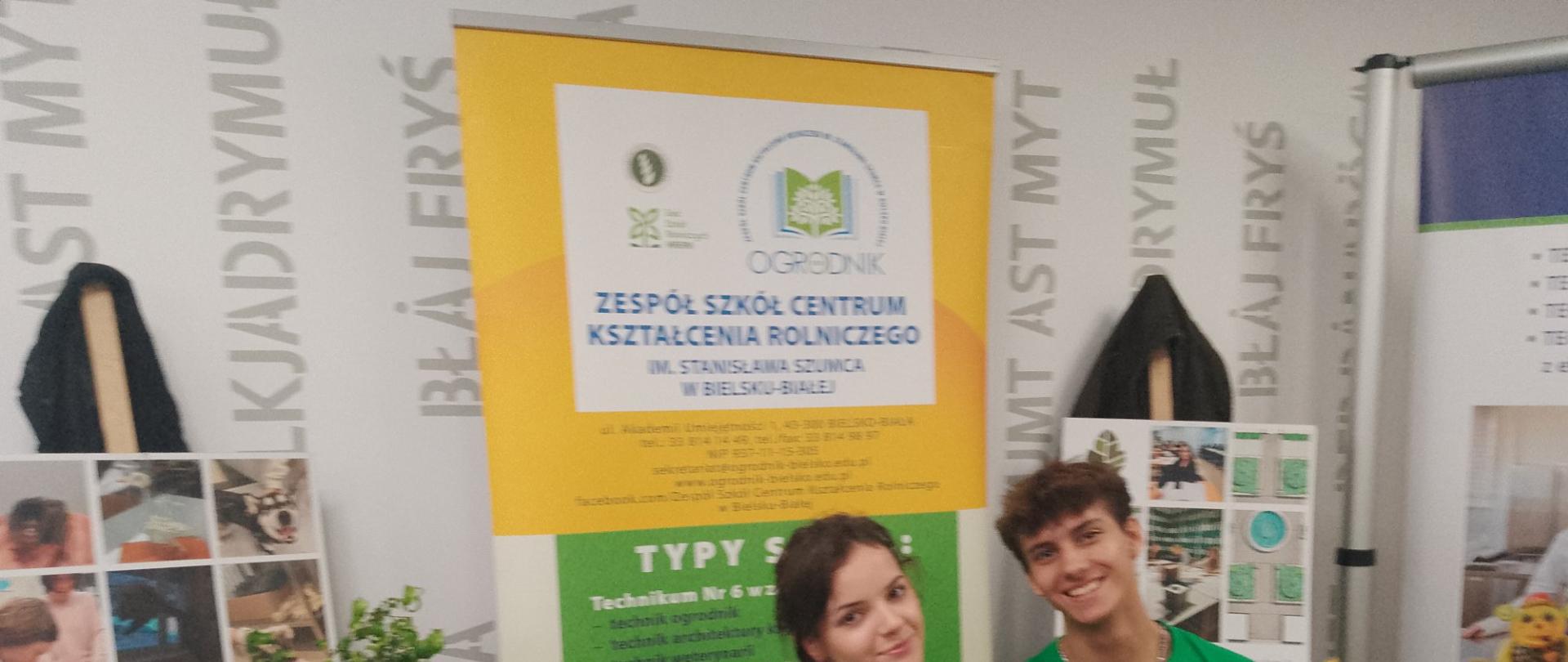 Dwie osoby siedzą przy stoisku ZSCKR; za nimi żółto‑zielony baner szkoły, na stole laptop z prezentacją, model budynku, kosz owoców i rośliny.