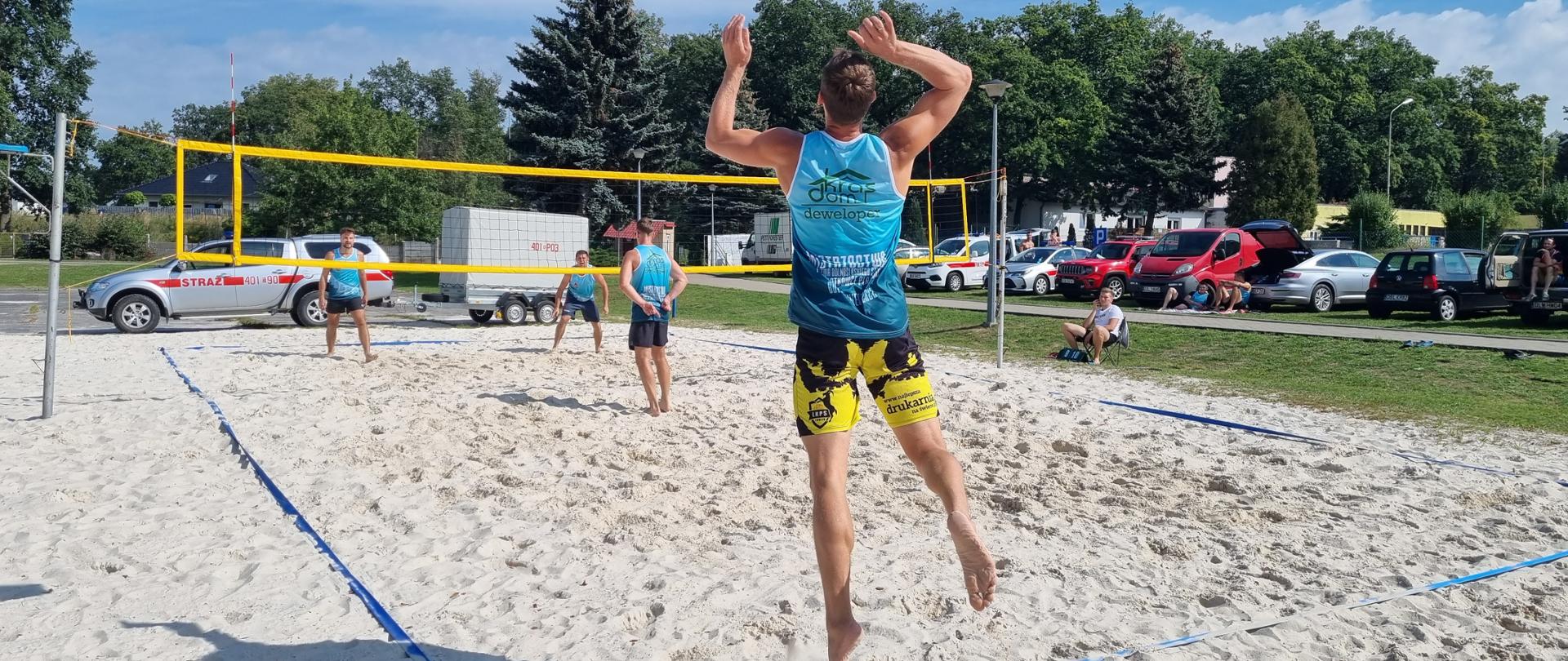 czterech zawodników w niebieskich koszulkach gra w siatkówkę plażową. W tle parking z samochodami 