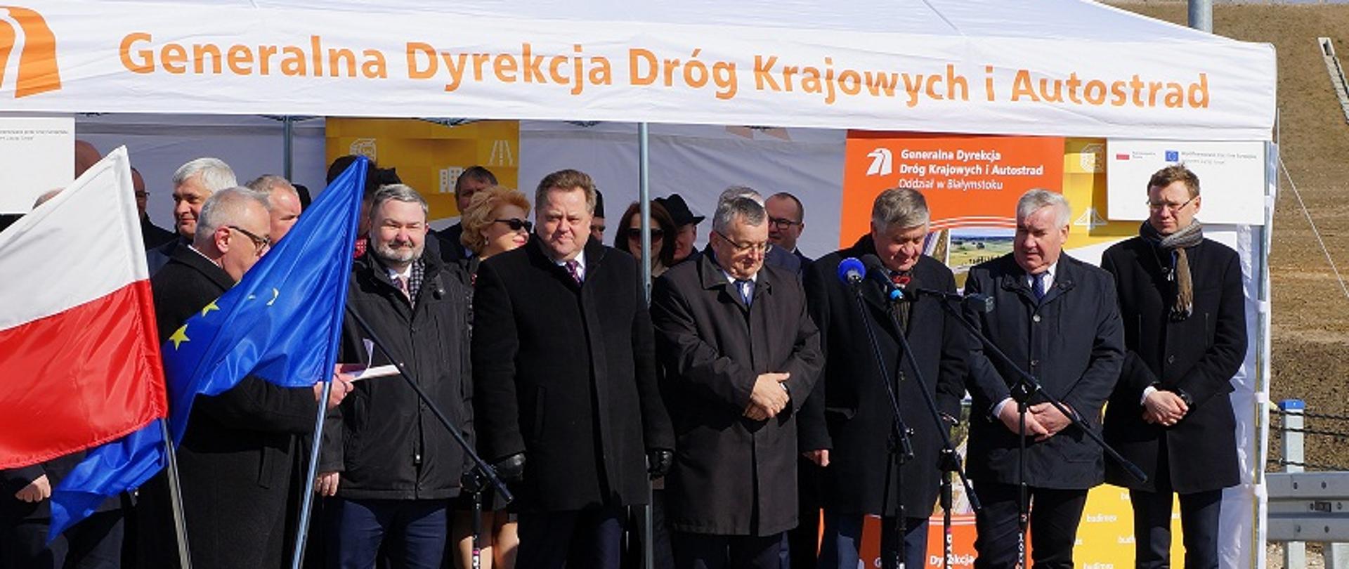 Na zdjęciu znajduję się grupa osób wśród nich między innymi Dyrektor Programów Infrastrukturalnych w Ministerstwie Inwestycji i Rozwoju Jarosław Orliński. Stoją pod białym namiotem z napisem "Generalna Dyrekcja Dróg Krajowych i Autostrad". Z boku stoi flaga Polski i Unii Europejskiej. 