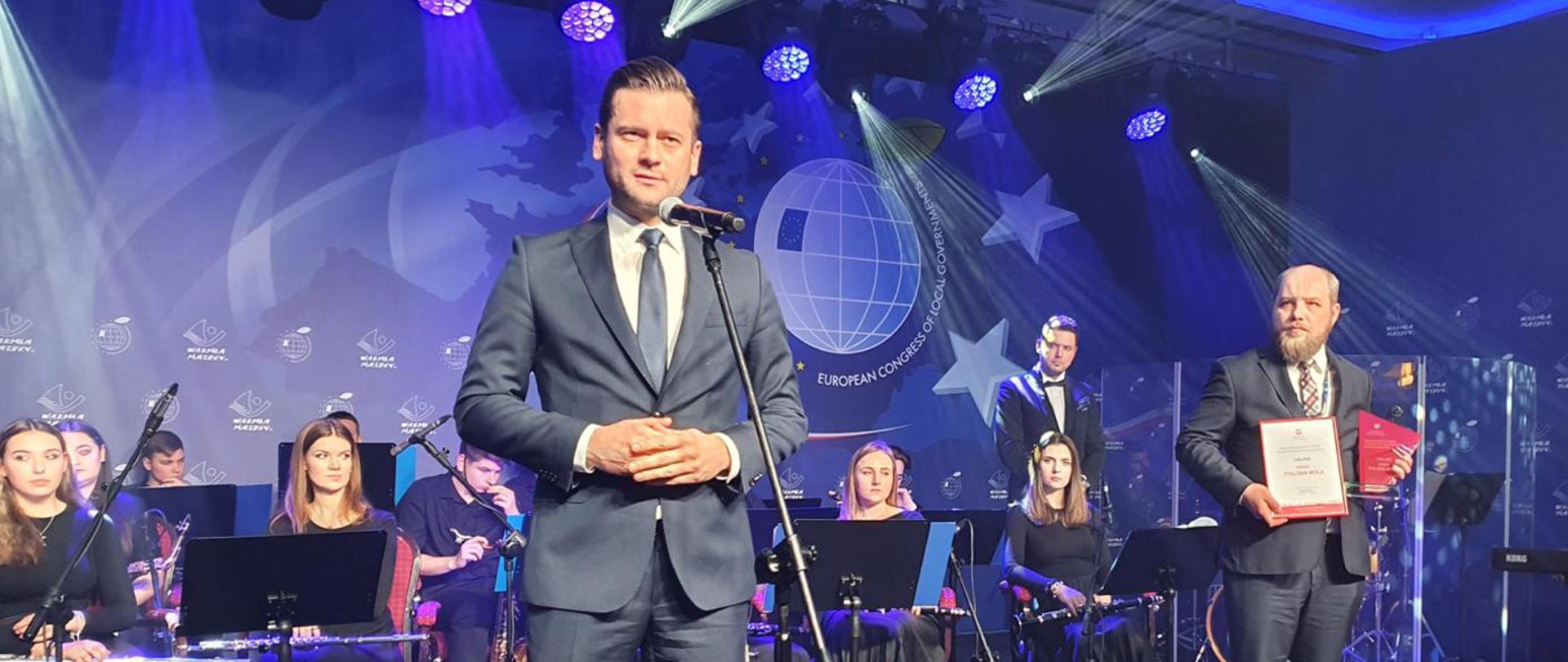 Minister Kamil Bortniczuk przemawia na Gali Nagród VII Europejskiego Kongresu Samorządowego. Minister stoi na scenie, mówi do mikrofonu. W tle na scenie widoczna orkiestra a także stojący z prawej strony przedstawiciel samorządu Miasto Stalowa Wola.