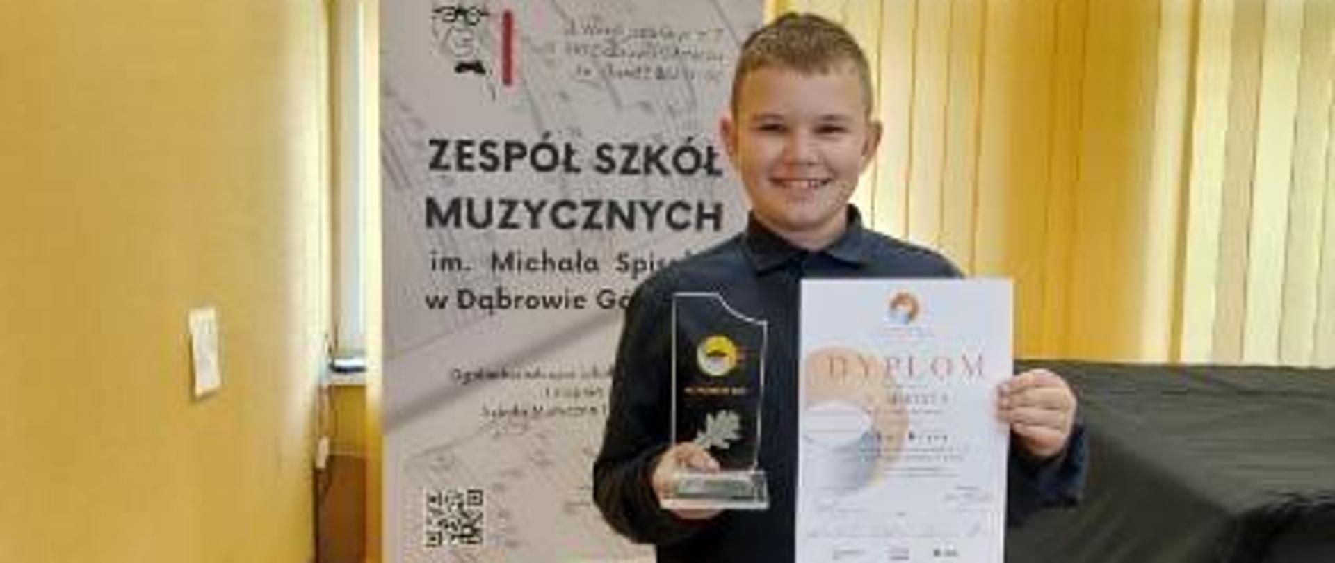 Na pierwszym planie stoi laureat konkursu z dyplomem i statuetką
w dłoni. Za nim ustawiony baner z nazwą instytucji: Zespół Szkół Muzycznych im. Michała Spisaka w Dąbrowie Górniczej.
