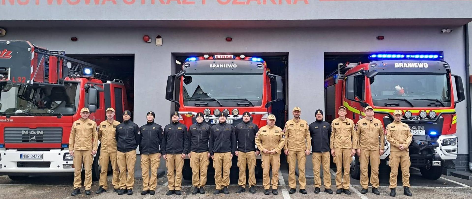 Na zdjęciu widzimy grupę strażaków ustawionych w szeregu przed budynkiem jednostki Państwowej Straży Pożarnej. Za nimi znajdują się trzy wozy strażackie.
Część strażaków ubrana jest w czarne kurtki i czapki zimowe, druga część w beżowe mundury polowe PSP z czapkami. Panuje porządek i dyscyplina – wszyscy stoją w równym rzędzie. Nad bramami garażowymi widnieje napis "PAŃSTWOWA STRAŻ POŻARNA". Całość tworzy wrażenie gotowości i profesjonalizmu
