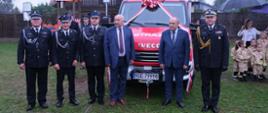 Zdjęcie zrobione na zewnątrz, podczas uroczystego poświęcenia i przekazania samochodu ratowniczo-gaśniczego dla Druhów ochotników. Na tle samochodu stoją zaproszenie goście wśród nich oficer Państwowej Straży Pożarnej w stopniu generała. 