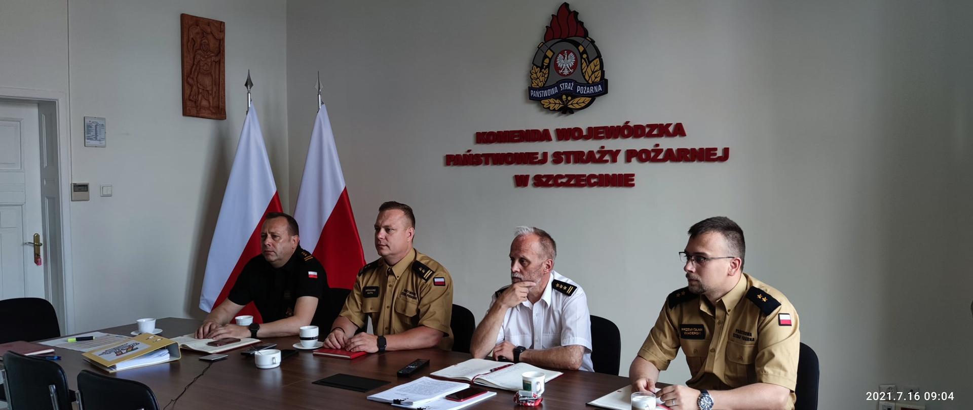 Funkcjonariusze Komendy Wojewódzkiej PSP w Szczecinie przy stole. W tle zdjęcia widoczne są flagi Polski, wraz z logiem PSP oraz drewniana ikona św. Floriana. 