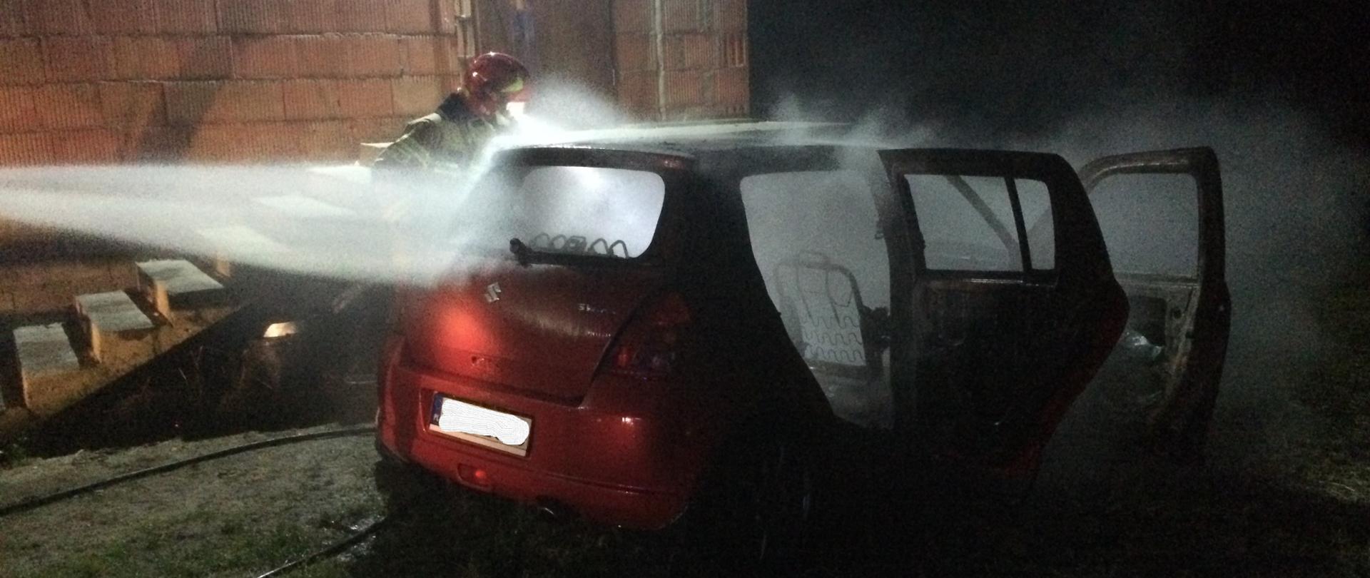 Samochód osobowy czerwony, który jest gaszony przez straż pożarną. Obok niego widać strażaka w stroju strażackim, a za i nim ścianę budynku z czerwonej cegły.