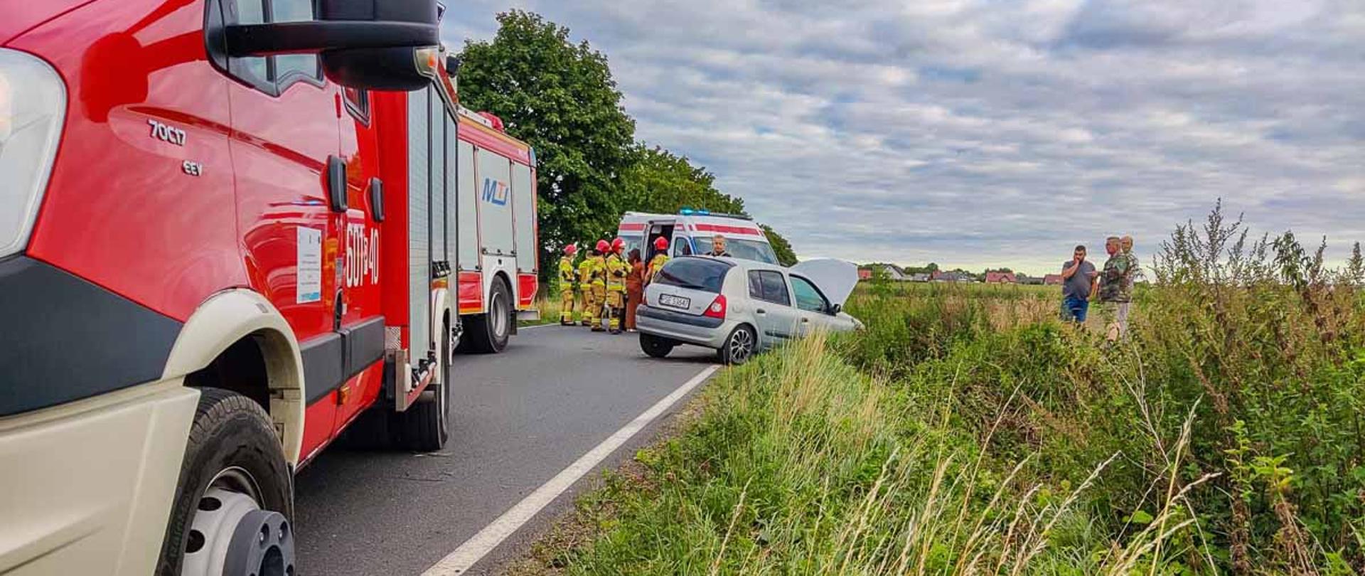 Na zdjęciu strażacy przy samochodach pożarniczych na drodze na poboczu w stronę rowu samochód osobowy za nim karetka pogotowia