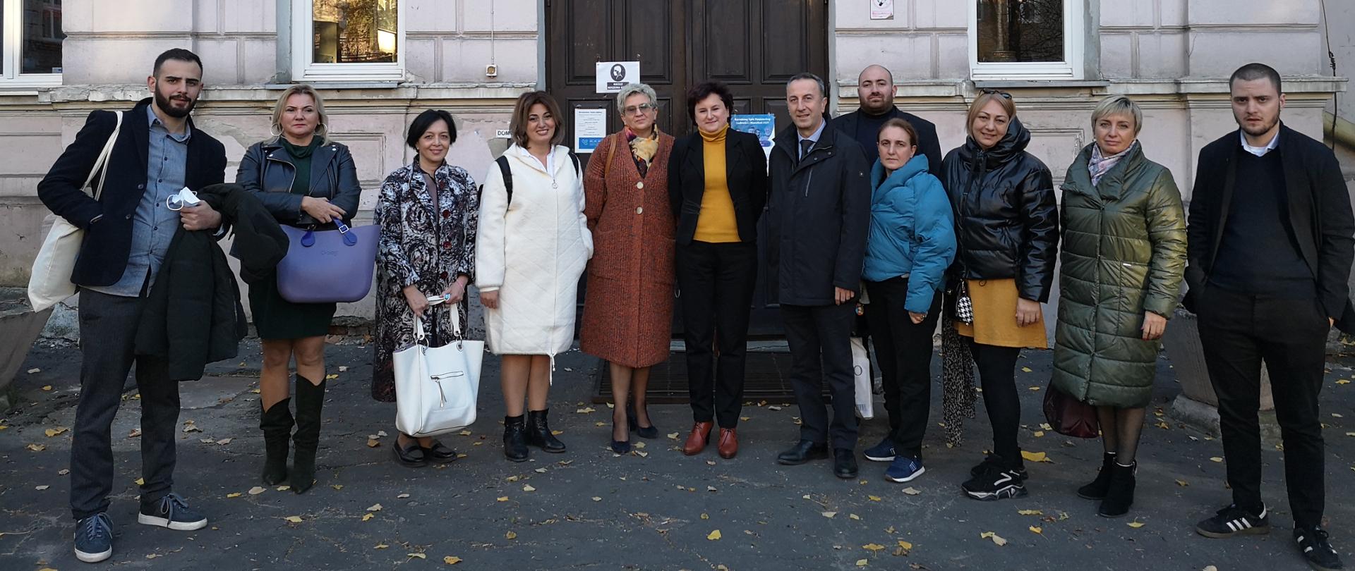 Dwanaście osób w jesiennych płaszczach stoi na tle zabytkowej kamienicy,.