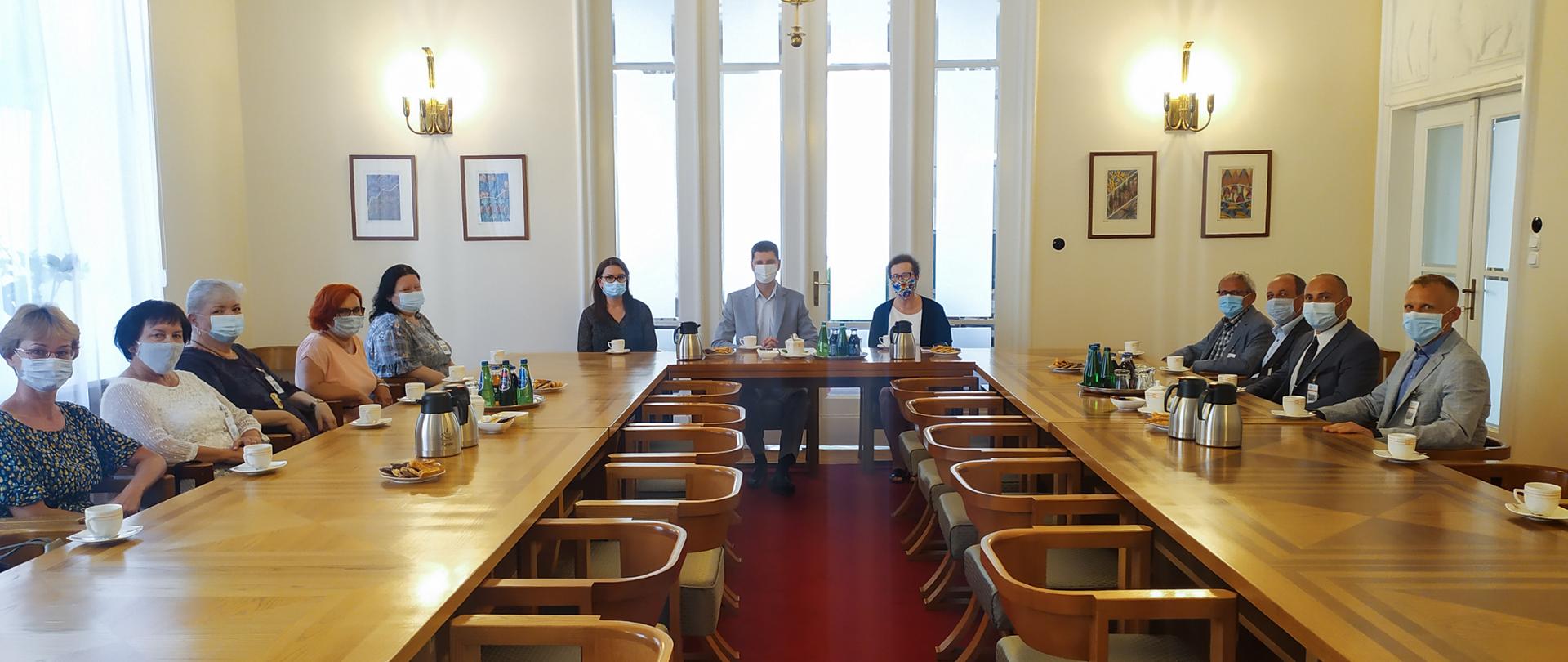 Uczestnicy spotkania siedzą przy stole w kształcie podkowy. W centrum siedzi wiceminister Dariusz Piontkowski.