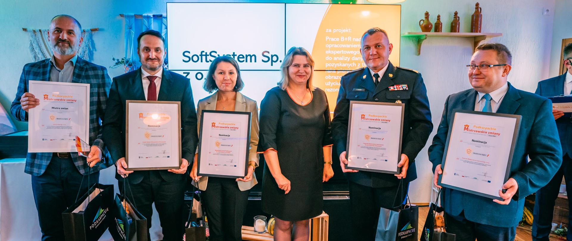 Zdjęcie wykonane w pomieszczeniu. Na pierwszym planie sześć osób stojących w szeregu. Pięć z nich trzyma oprawione w ramki dyplomy. Wśród nich funkcjonariusz Państwowej Straży Pożarnej w stopniu generała, w mundurze wyjściowym.