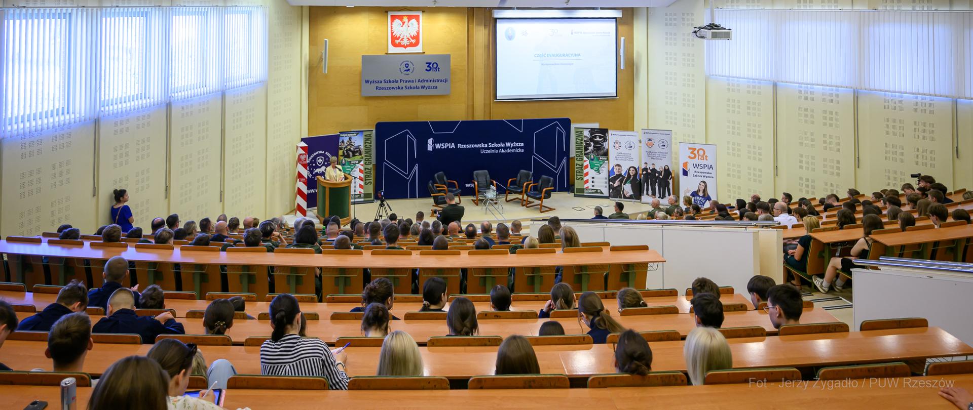 W sali WSPiA Rzeszowskiej Szkoły Wyższej siedzą licznie przybyli uczestnicy konferencji. Przy mównicy stoi Wojewoda Podkarpacki. 