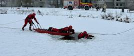 Strażacy w czerwonych kombinezonach do ratownictwa wodnego ćwiczą wydobywanie osoby z przerębla przy użyciu specjalnych sani lodowych. W tle widoczny jest wóz strażacki oraz nowo powstające budynki mieszkalne w zimowej scenerii.