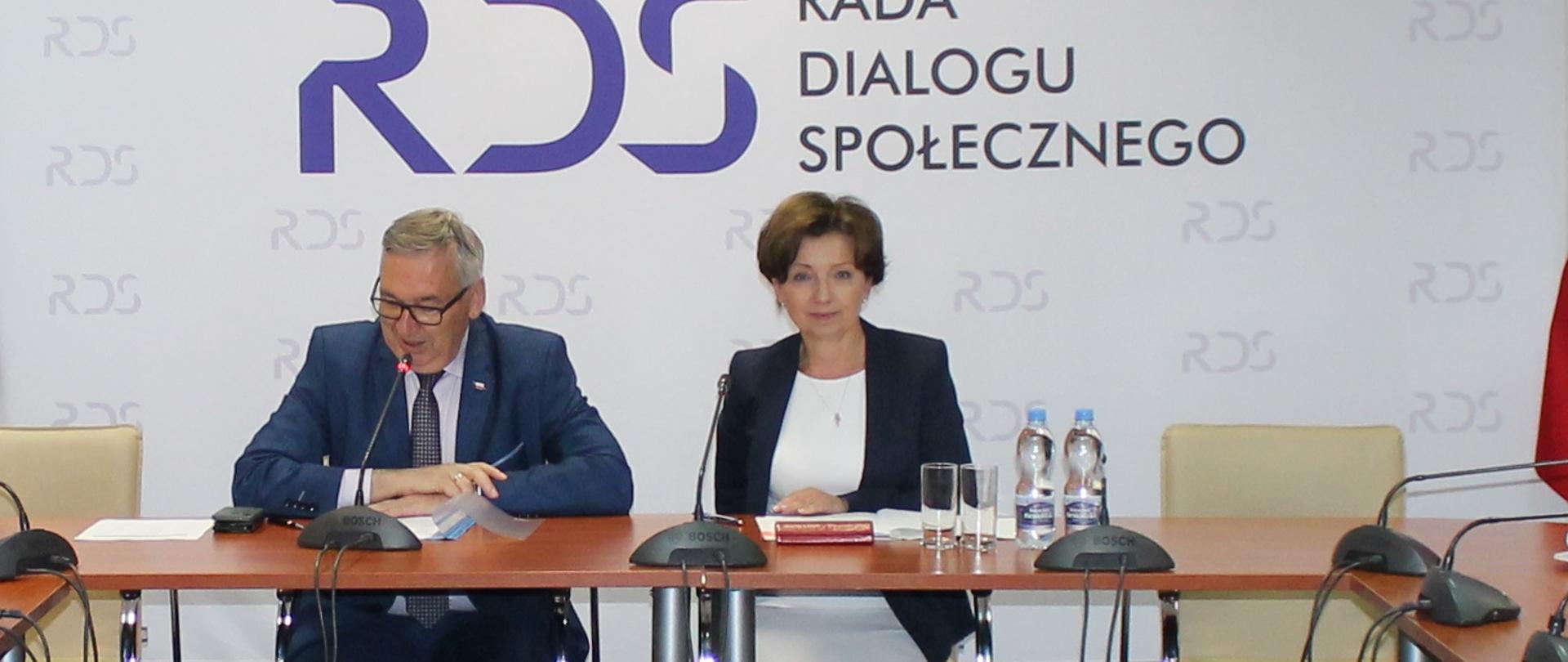 Na zdjęciu znajduje się stół prezydialny za którym siedzą od lewej strony: Stanisław Szwed - Sekretarz Stanu w Ministerstwie Rodziny i Polityki Społecznej a następnie Marlena Maląg Minister Rodziny i Polityki Społecznej. W tle siedzących osób znajduje się baner z napisem Rada Dialogu Społecznego. Każdej z osób przypisany jest mikrofon.
