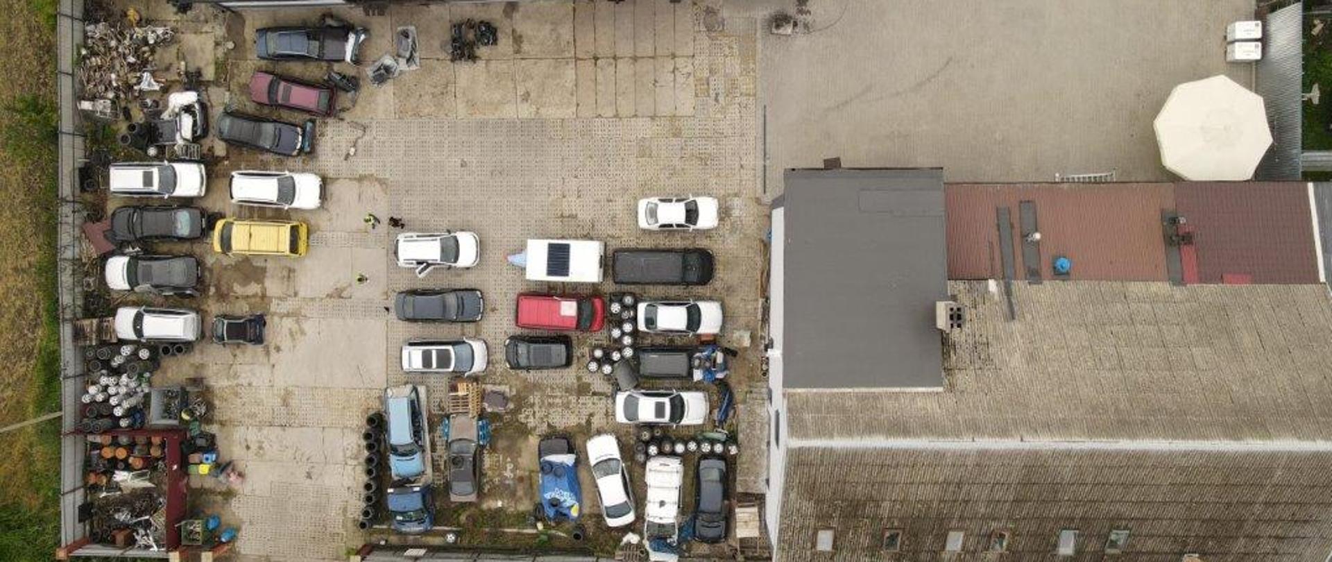 Widok z drona na miejsce nielegalnego demontażu. Widoczne pojazdy w różnym stopniu poddane demontażowi, części, opony oraz pojemniki z odpadami.