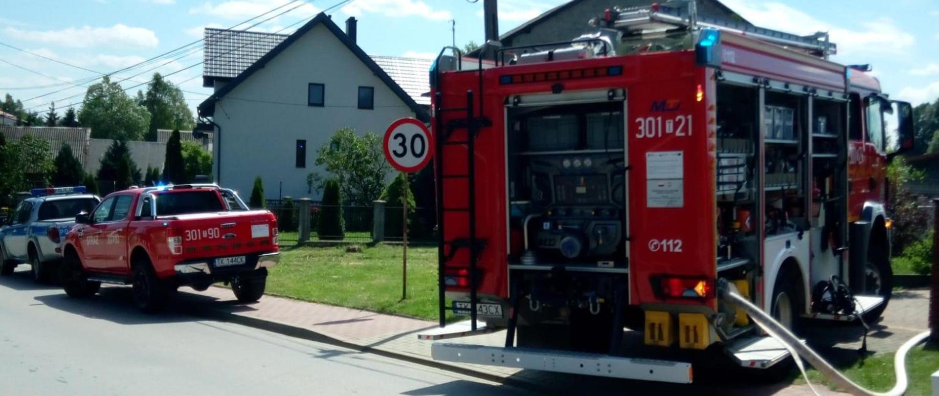 Zdjęcie przedstawia drogę gdzie zaparkowały dwa samochody pożarnicze oraz radiowóz policyjny biorące w akcji gaśniczej w Mójczy.