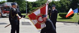 po lewej strażacy w poczcie sztandarowym, po lewej zdający obowiązki bryg. Radosław Lendor Komendant Miejski Państwowej Straży Pożarnej w Sosnowcu klęczący przed sztandarem komendy, obok mł. bryg. Marcin Pilarski przejmujący obowiązki Komendanta Miejskiego Państwowej Straży Pożarnej w Sosnowcu
