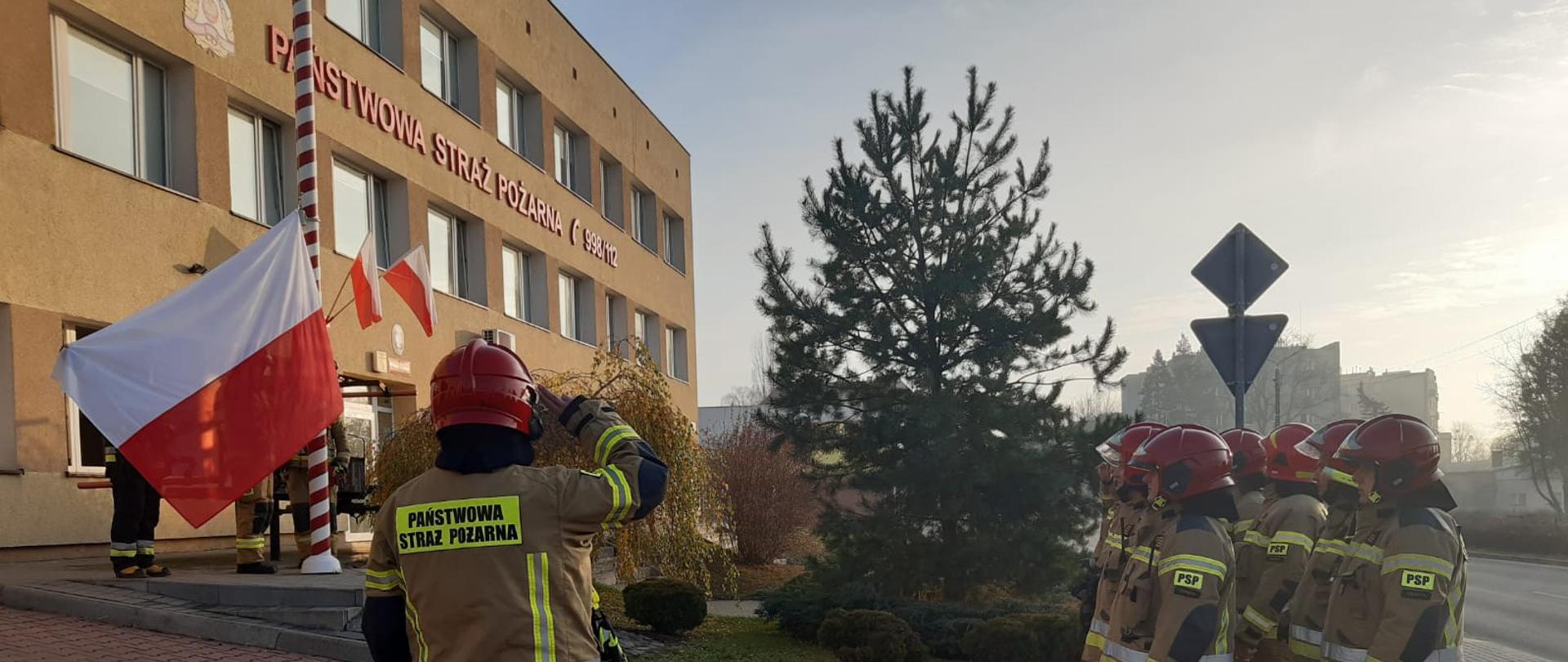 Na zdjęciu widać strażaków ubranych w ubranie specjalne koloru piaskowego w czerwonych hełmach, ustawionych w dwuszeregu przed masztem na który wciągana jest biało-czerwona flaga.