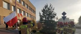 Na zdjęciu widać strażaków ubranych w ubranie specjalne koloru piaskowego w czerwonych hełmach, ustawionych w dwuszeregu przed masztem na który wciągana jest biało-czerwona flaga.