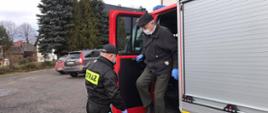 Osoba starsza – mężczyzna wysiada z samochodu pożarniczego koloru czerwonego Ochotniczej Straży Pożarnej w Trzcianie przez otwarte drzwi z tyłu samochodu. Mężczyzna zabezpieczony jest dodatkowo w maseczkę oraz rękawiczki lateksowe. Osoba wysiadająca korzysta z pomocy podnóżka podczas wysiadania. Strażak OSP ubrany w czarne ubranie koszarowe przytrzymuje drzwi wejściowe do pojazdu. Samochód stoi na parkingu w pobliżu ośrodka zdrowia