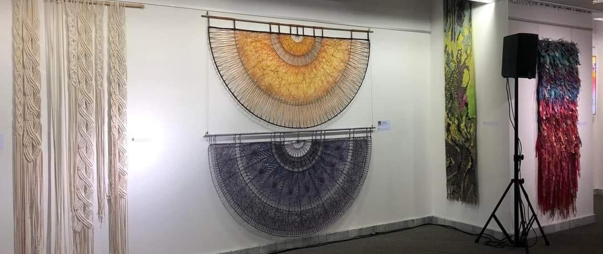 Prace z tkaniny artystycznej powieszone na ścianie w galerii. W środku dwie nagrodzone prace A. Kwiecień. Przypominają one olbrzymie wachlarze. Jeden jest koloru fioletowego, czarnego,, niebieskiego Drugi jest w kolorach żółty i beżowy. Przypominają sieć.