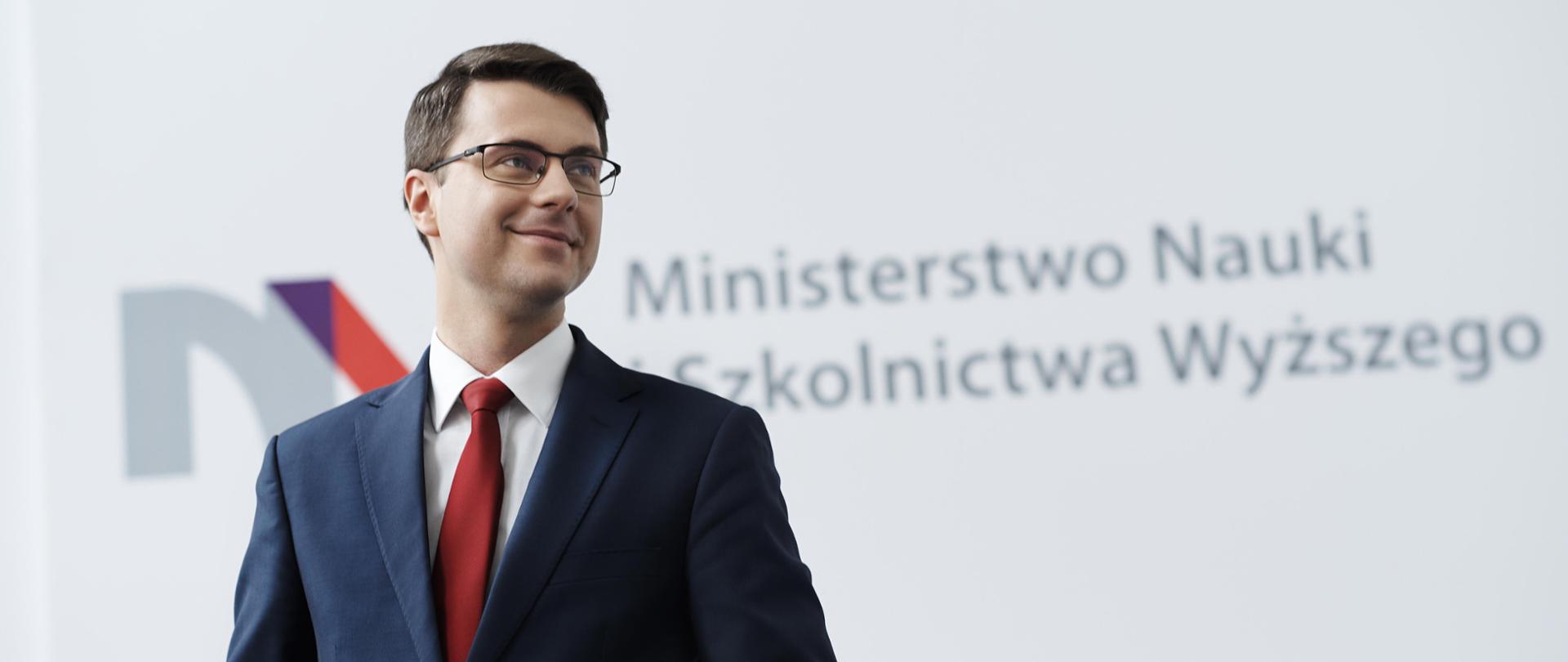 Wiceminister Piotr Müller stoi na tle białej ściany z logiem MNiSW