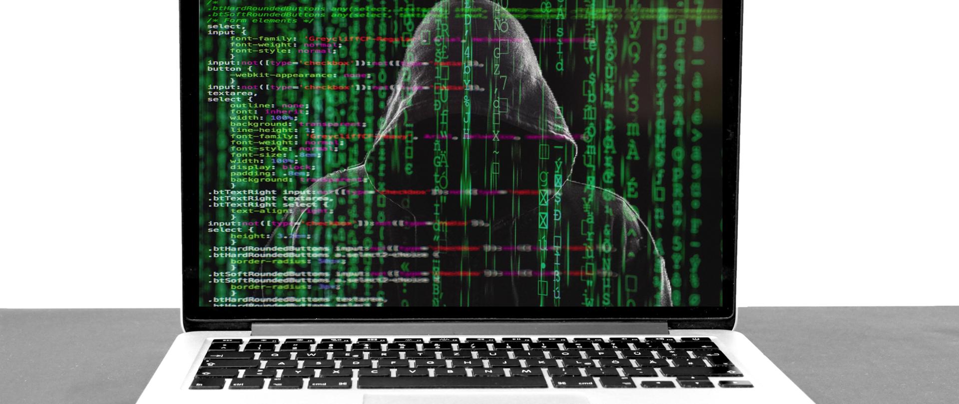 Zdjęcie laptopa. Na ekranie laptopa zamaskowana osoba. W tle kody systemowe.