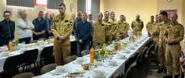 Funkcjonariusze, pracownicy cywili oraz emeryci stoją przy zastawionych potrawami i napojami stołach słuchając składanych życzeń z okazji Świąt Wielkanocnych.