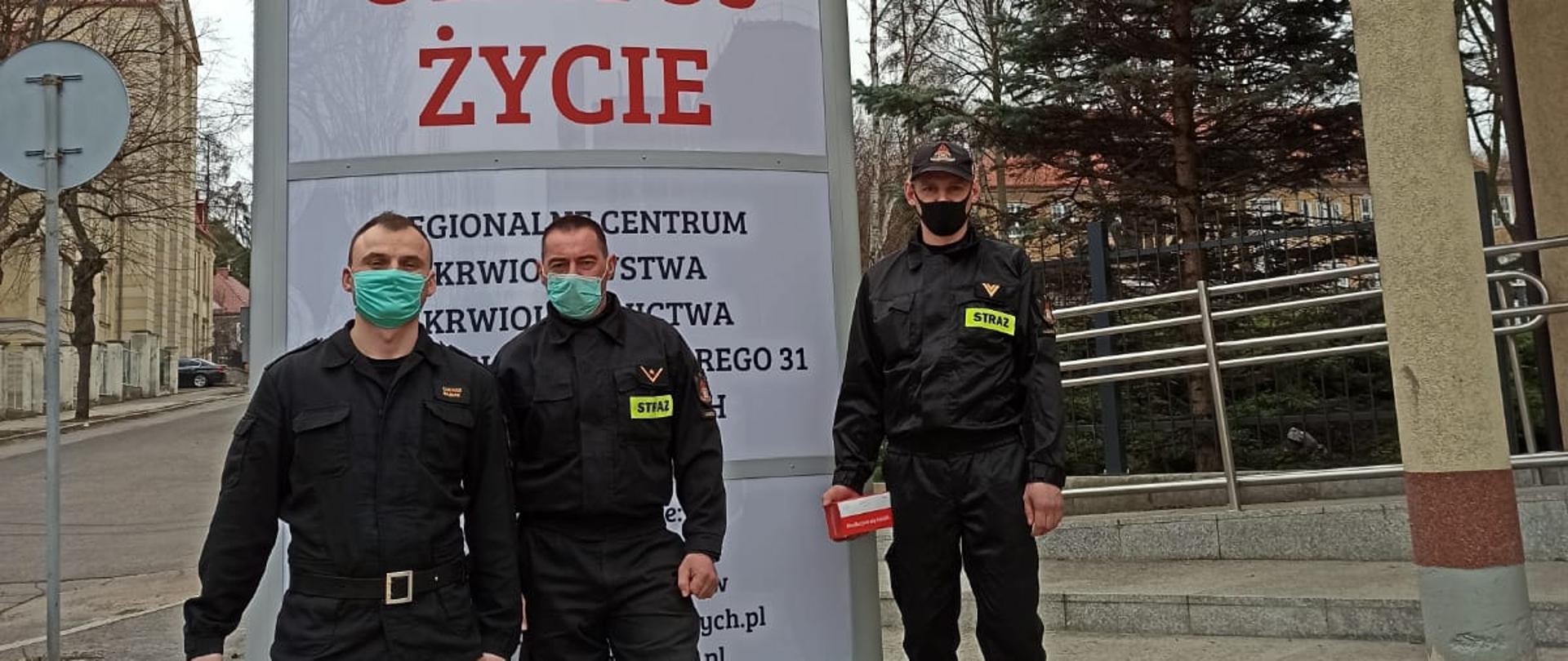 Na pierwszym planie trzech funkcjonariuszy w umundurowaniu bojowym z założonymi na twarzy maseczkami. Za nimi pylon reklamowy Regionalnego Centrum Krwiodawstwa i Krwiolecznictwa w Wałbrzychu.