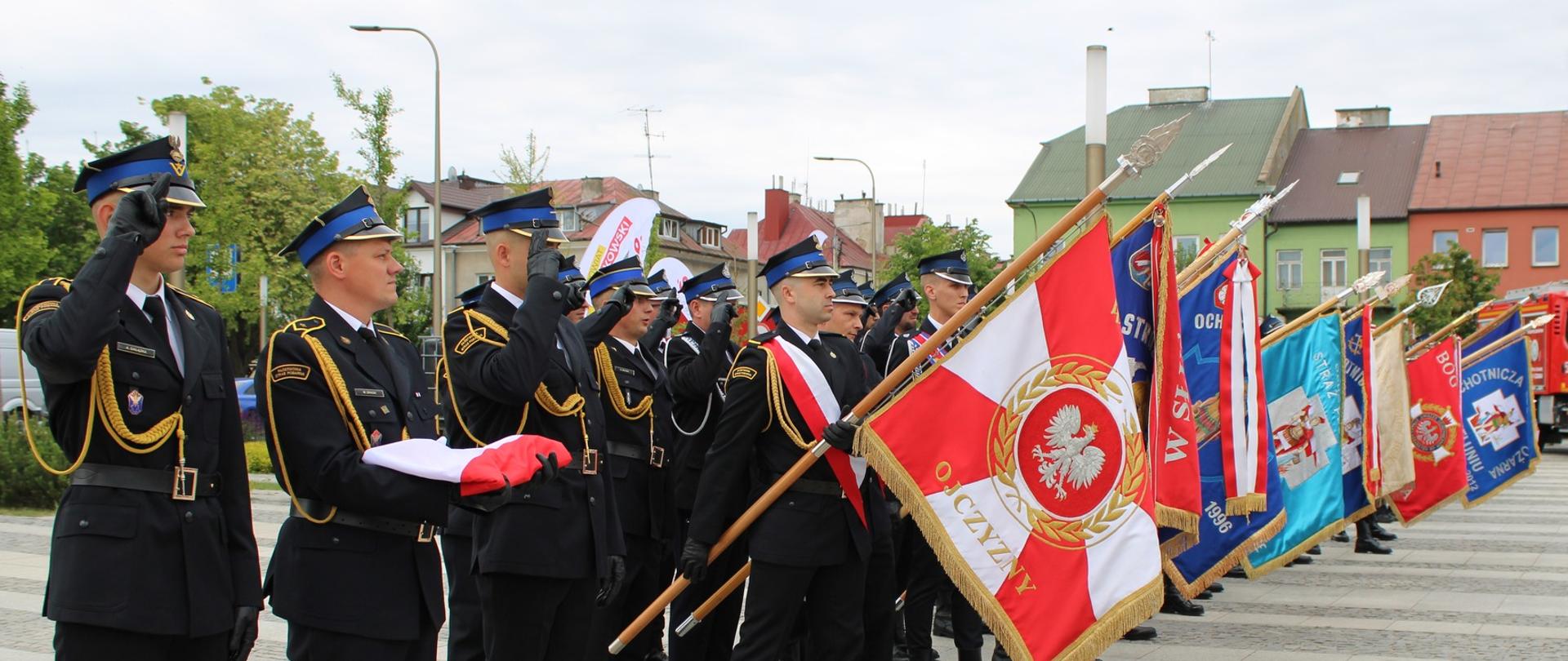 Poczet flagowy oraz liczne poczty sztandarowe stoją w rzędzie na placu podczas apelu. Na pierwszym planie trzej strażacy trzymają złożoną polską flagę państwową, a dwaj z nich salutują. Za nimi stoją kolejni mundurowi z pochylonymi sztandarami jednostek OSP i PSP, z których najbliższy ma czerwono-białe barwy z godłem Polski i napisem „OJCZYŹNIE”. W tle widoczny jest wóz strażacki i zabudowania miejskie.