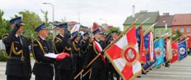 Poczet flagowy oraz liczne poczty sztandarowe stoją w rzędzie na placu podczas apelu. Na pierwszym planie trzej strażacy trzymają złożoną polską flagę państwową, a dwaj z nich salutują. Za nimi stoją kolejni mundurowi z pochylonymi sztandarami jednostek OSP i PSP, z których najbliższy ma czerwono-białe barwy z godłem Polski i napisem „OJCZYŹNIE”. W tle widoczny jest wóz strażacki i zabudowania miejskie.