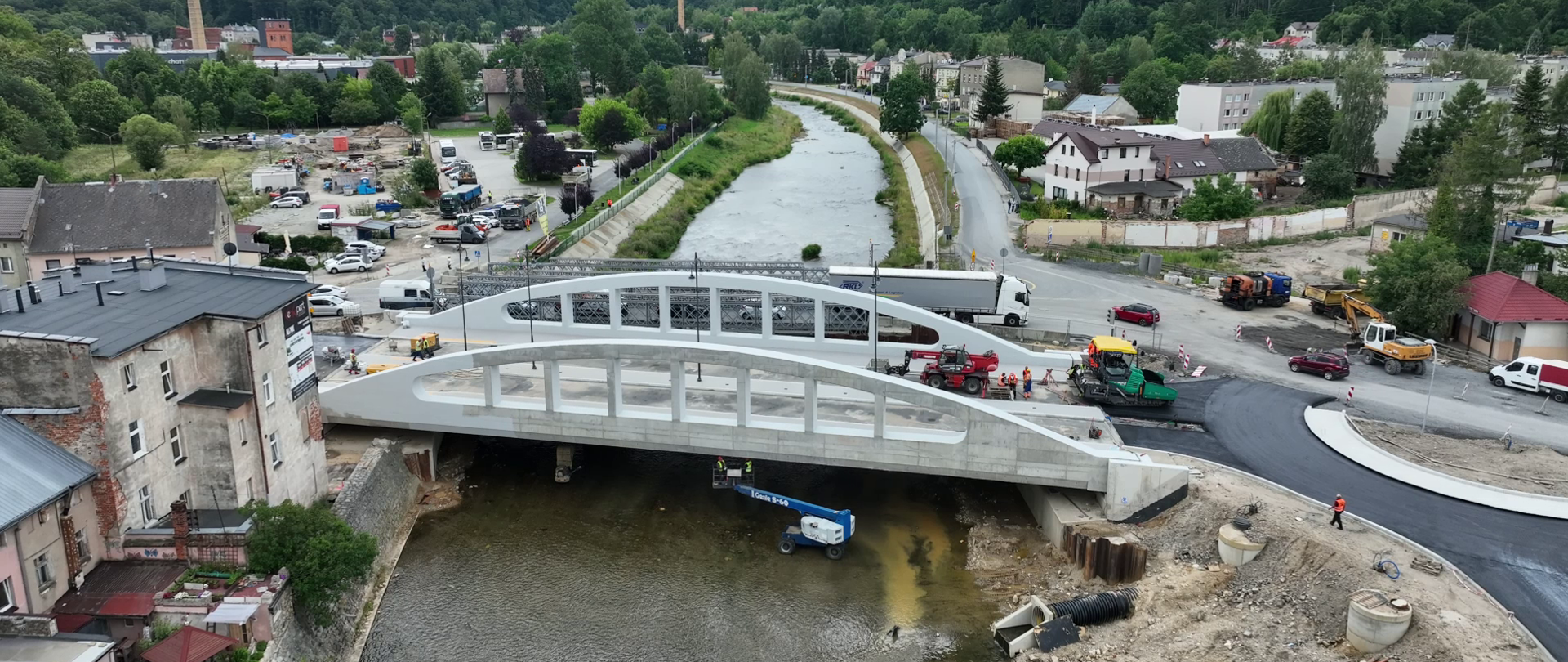 Widok z lotu ptaka na nowy most nad rzeką w trakcie realizacji inwestycji drogowej. Na moście i dojazdach pracują maszyny budowlane oraz ekipy drogowe. Wokół widoczne są zabudowania miejscowości, biegnąca wzdłuż rzeki droga oraz zalesione wzgórza w tle pod zachmurzonym niebem.