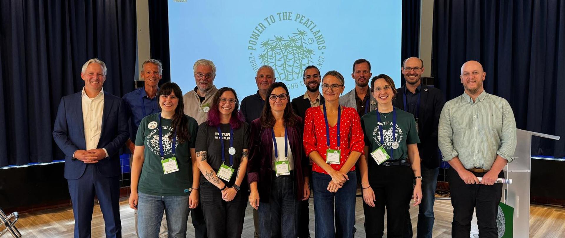 Na zdjęciu widoczna jest grupa uczestników stojących do wspólnego zdjęcia w sali konferencyjnej. W tle znajduje się ekran z wyświetlonym logo i napisem „Power to the Peatlands”, część osób ma identyfikatory konferencyjne. Wnętrze jest nowoczesne, z ciemnymi zasłonami i drewnianą podłogą, co nadaje wydarzeniu oficjalny charakter.
