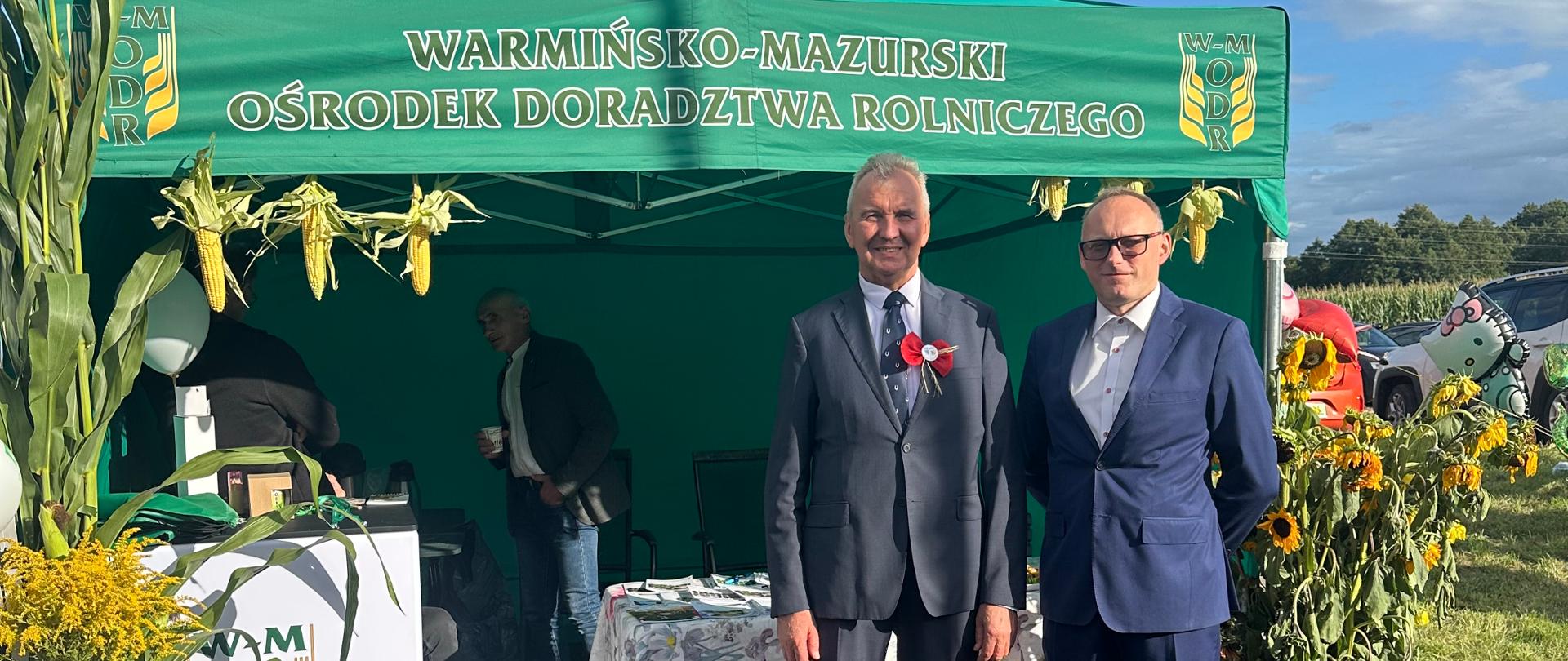 Dwóch mężczyzn stojących przed zielonym namiotem Warmińsko – Mazurskiego Ośrodka Doradztwa Rolniczego podczas imprezy plenerowej