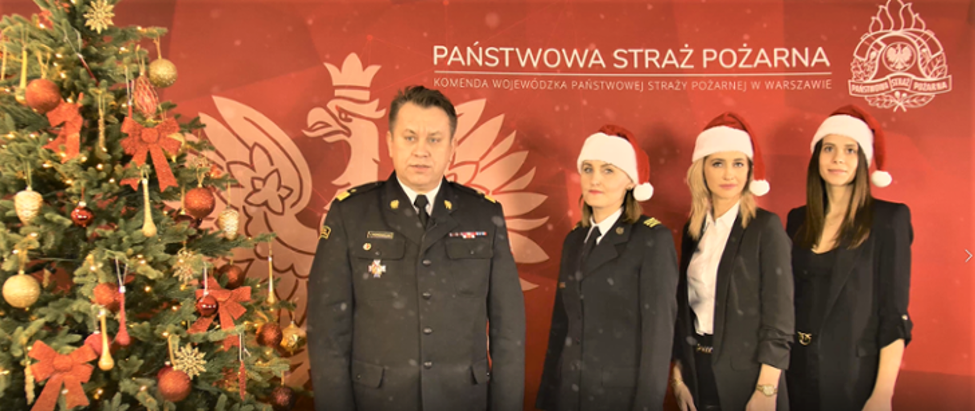 Na zdjęciu widoczny komendant wojewódzki PSP w mundurze wyjściowym. Po jego prawej stronie ubrana, świąteczna choinka, a po lewej trzy kobiety - jedna w mundurze wyjściowym, pozostałe dwie w ubraniach cywilnych, z czapkami mikołaja na głowach. W tle czerwona plansza z orłem, logiem PSP i napisem Państwowa Straż Pożarna.
