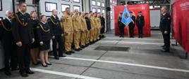 Strażacy stoją ustawieni w dwuszeregu w mundurach czarnch i zółtych, poczet sztandarwy stoi na tle ścianki firmowej. Widoczne bramuy garażowe.