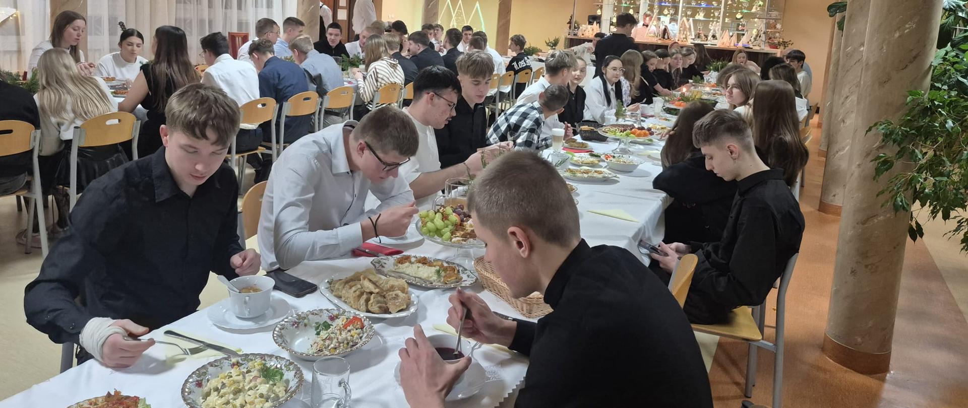 Za dwoma długimi wigilijnymi stołami siedzą mieszkańcy internatu. Rozmawiają i jedzą świąteczne potrawy.