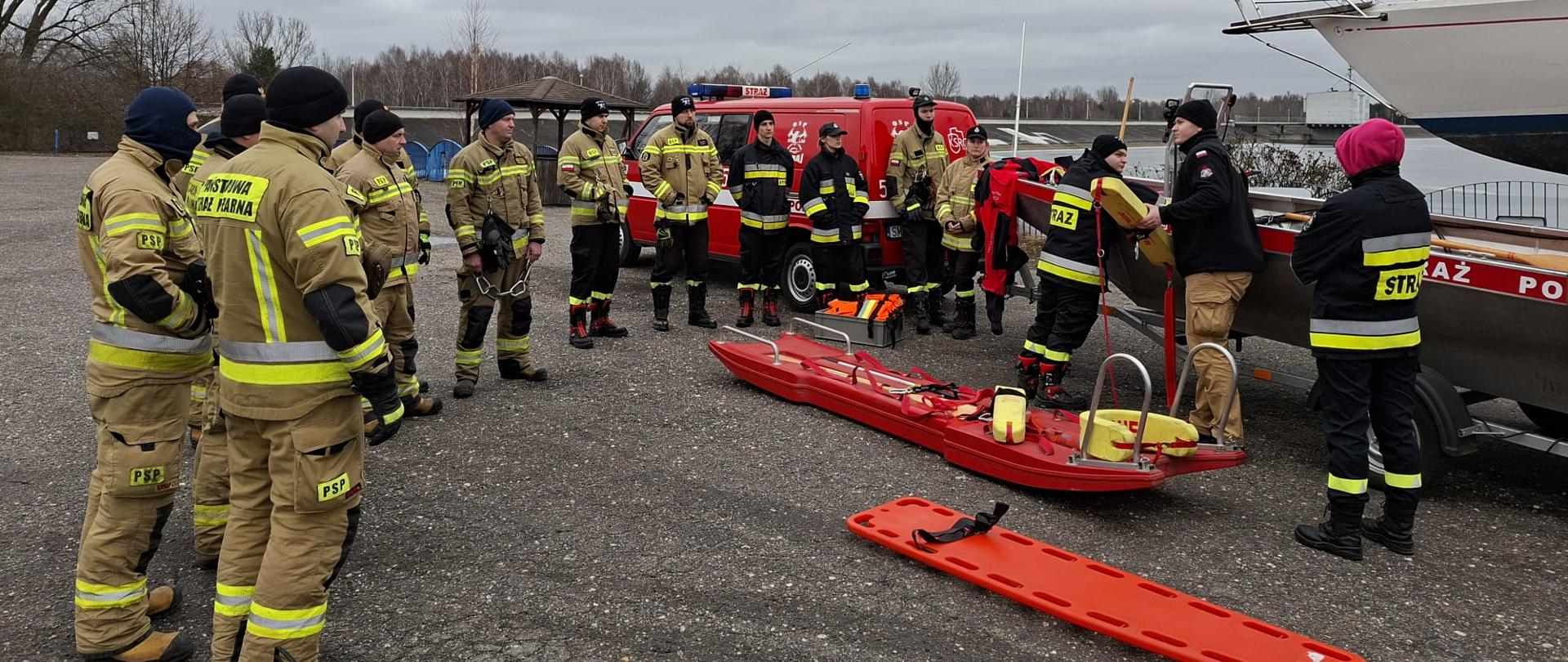 Grupa kilkunastu strażaków w ubraniach specjalnych stoi na brzegu zbiornika wodnego podczas szkolenia z zakresu działań ratowniczych na lodzie. Instruktor omawia zasady użycia sprzętu ratowniczego przy łodzi, obok której na ziemi leżą czerwone sanie lodowe oraz deska ortopedyczna. W tle widoczny jest samochód ratowniczy, całość odbywa się w pochmurny, zimowy dzień.