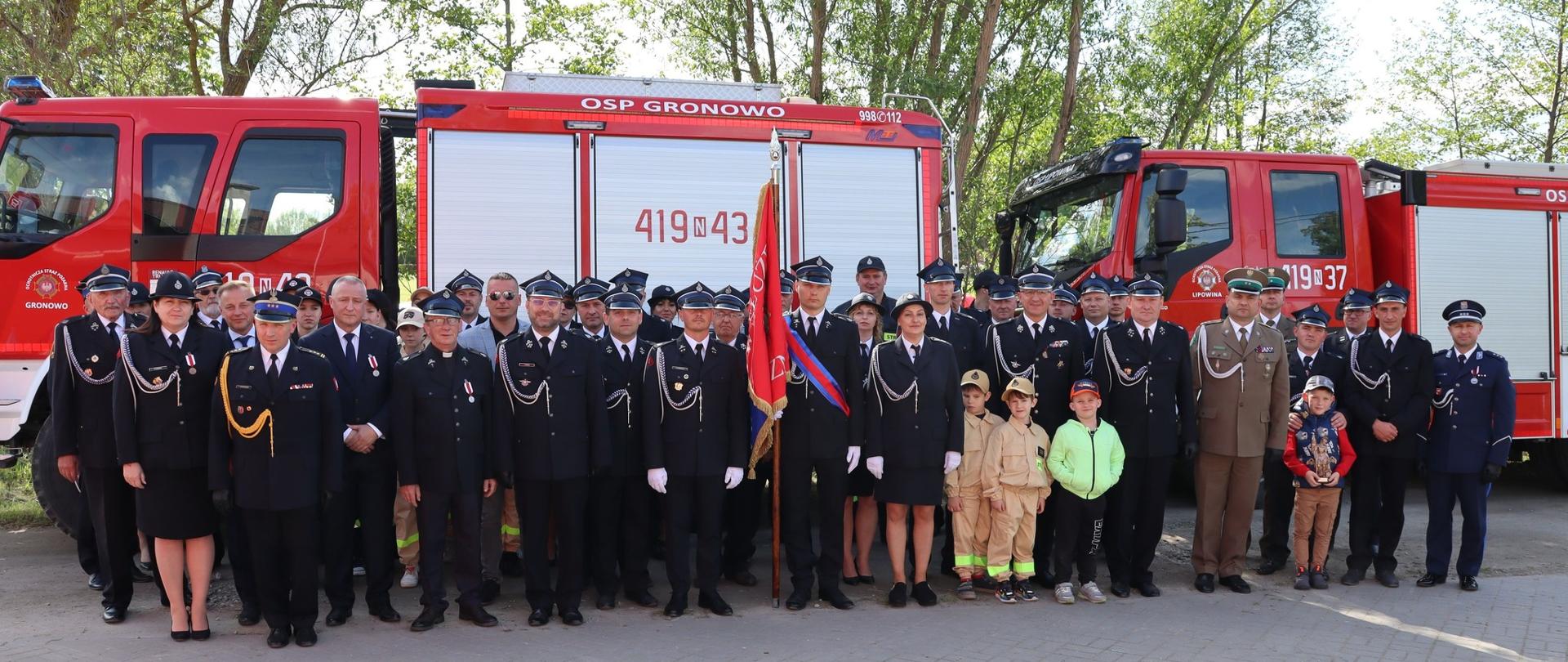 Duża grupa strażaków w mundurach galowych i zaproszonych gości, stoją na tle dwóch samochodów strażackich.