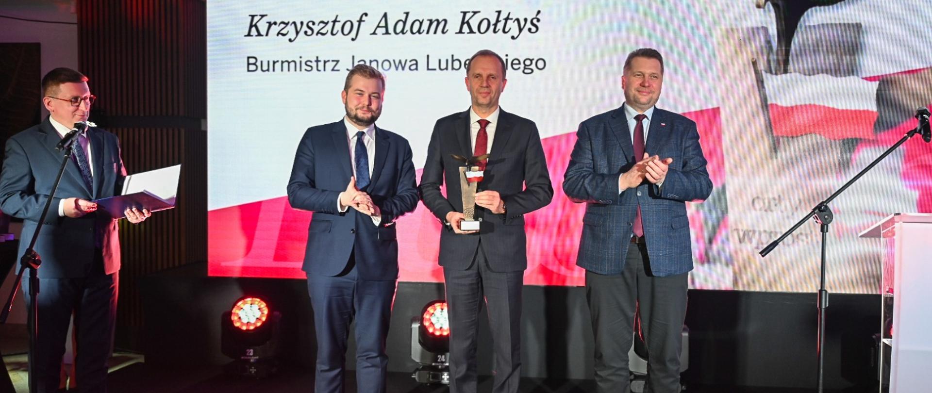 Minister Czarnek i trzech mężczyzn w garniturach stoją pod biało-czerwoną ścianką z napisem Krzysztof Adam Kołtyś i rysunkiem lecącego orła.