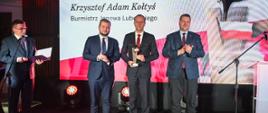 Minister Czarnek i trzech mężczyzn w garniturach stoją pod biało-czerwoną ścianką z napisem Krzysztof Adam Kołtyś i rysunkiem lecącego orła.
