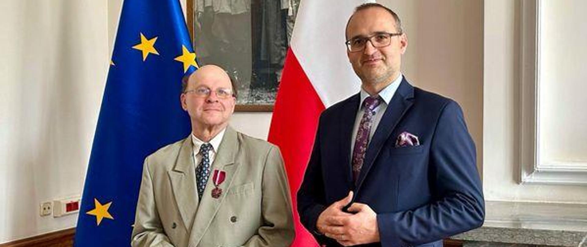 dwóch mężczyzn stoi na tle flag polskiej i unijnej. Jeden ma w klapie medal Gloria artis