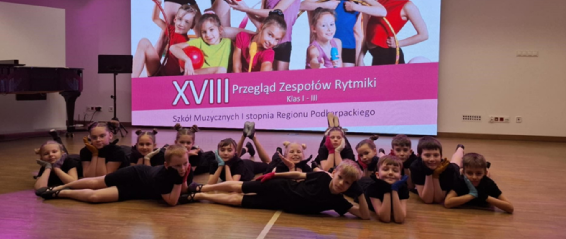 Grupa dzieci w czarnych strojach gimnastycznych leży na parkiecie w sali, uśmiechając się do aparatu. W tle widoczny jest duży ekran z kolorowym plakatem promującym XVIII Przegląd Zespołów Rytmiki dla klas II–III Szkół Muzycznych I stopnia Regionu Podkarpackiego. Plakat przedstawia dzieci w strojach do ćwiczeń rytmicznych.