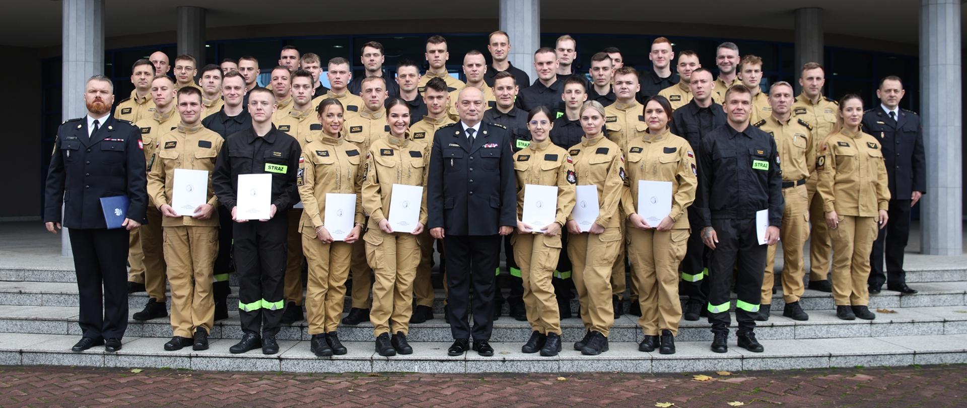 Pamiątkowe zdjęcie grupowe funkcjonariuszy i funkcjonariuszek będących uczestnikami czwartego turnusu Szkolenia Podstawowego w Zawodzie Strażak (37 osób) w towarzystwie Komendanta Centralnej Szkoły PSP, funkcjonariuszki CS PSP oraz siedmiu funkcjonariuszy CS PSP